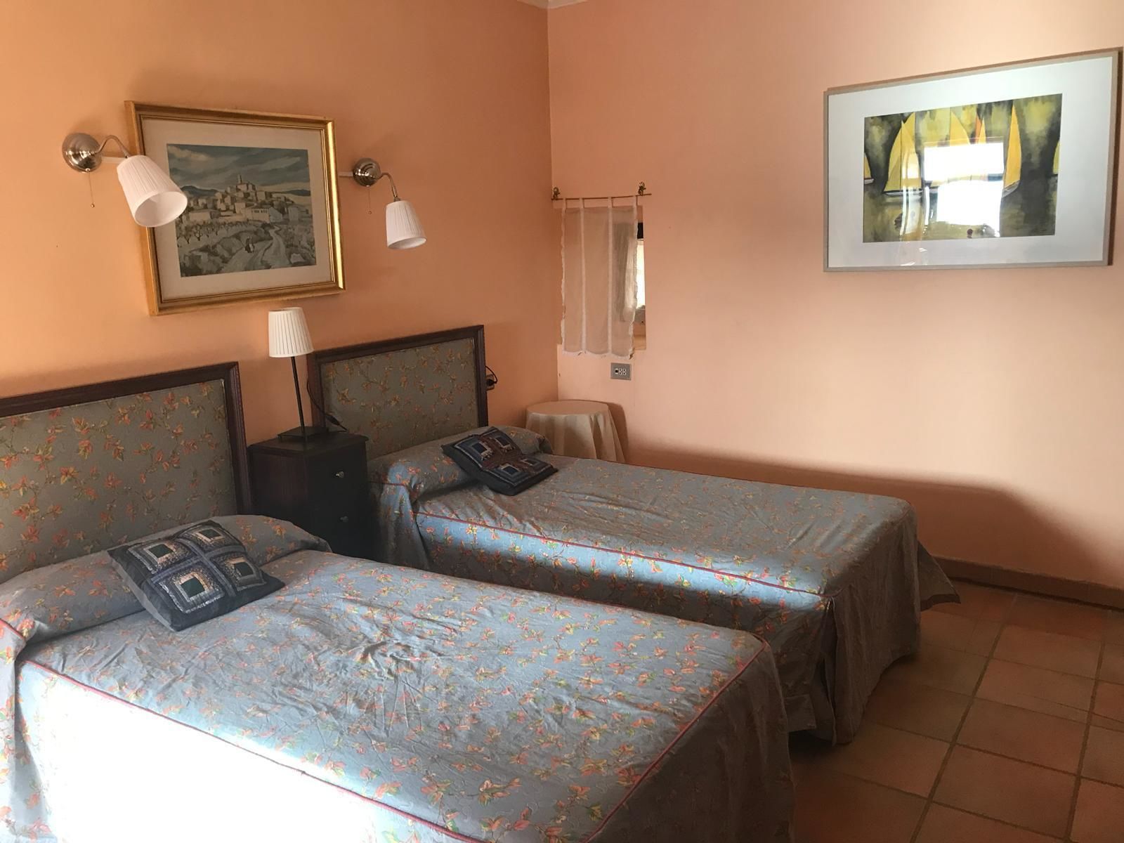 Habitació d'hotel amb dos llits, una tauleta de nit, una làmpada, un quadre amb marc i una finestra amb cortines.