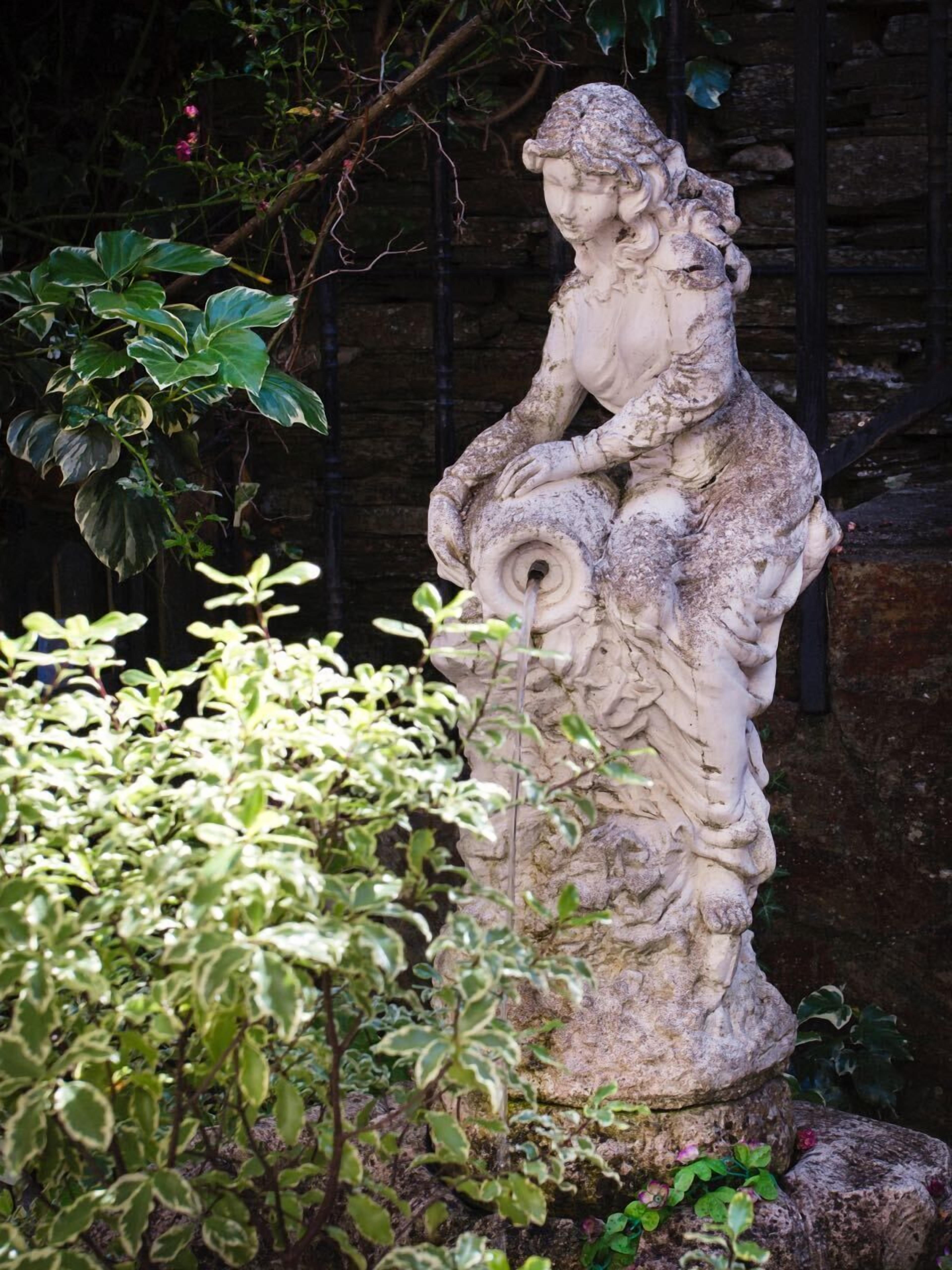 A stone statue of a woman holding a scroll, surrounded by greenery and a stone wall.