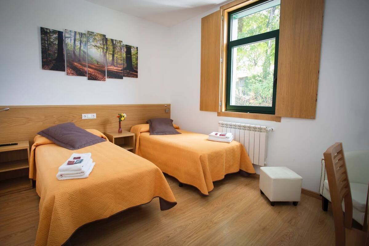 A room with two beds, a wooden headboard, a window with a view of trees, and a radiator.