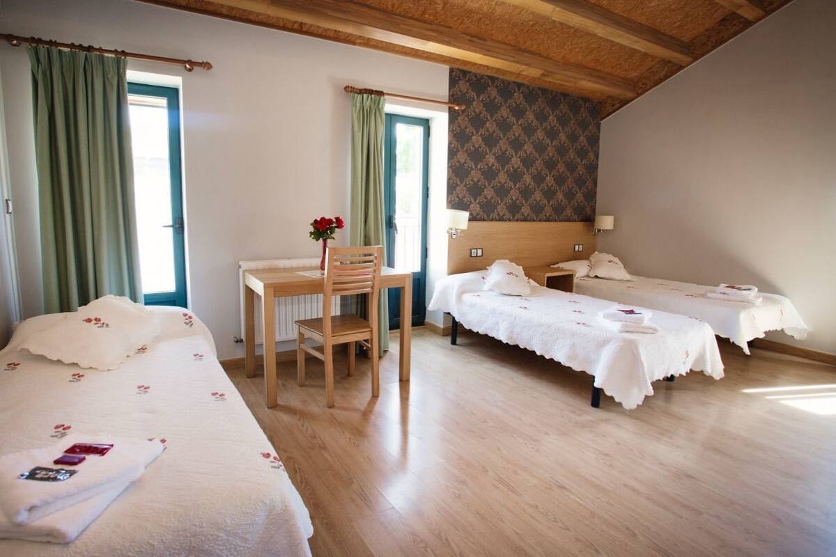 A hotel room with two single beds, a wooden desk, a chair, and a small table with a vase of flowers.