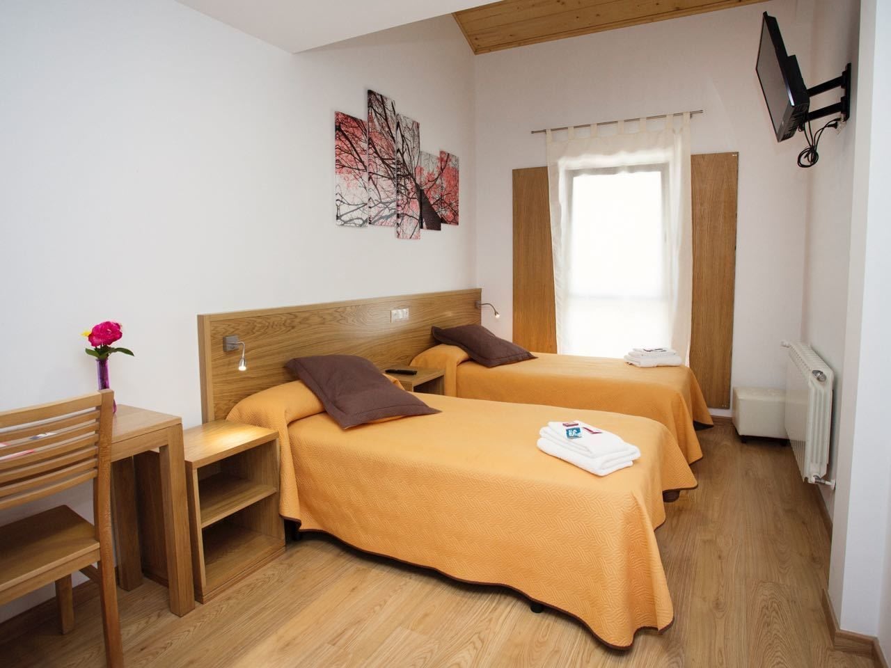 A hotel room with two beds, a wooden desk, a chair, a small table, a painting on the wall, a window with curtains, and a television mounted on the wall.