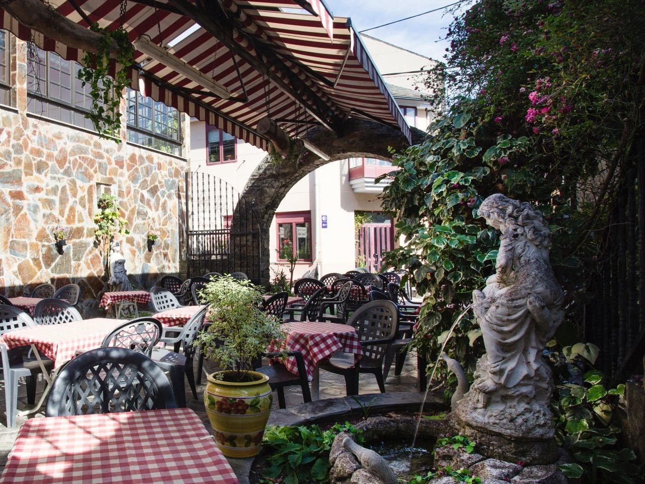 Patio esterno con pareti in pietra, tavoli a scacchi rossi e bianchi e una statua.