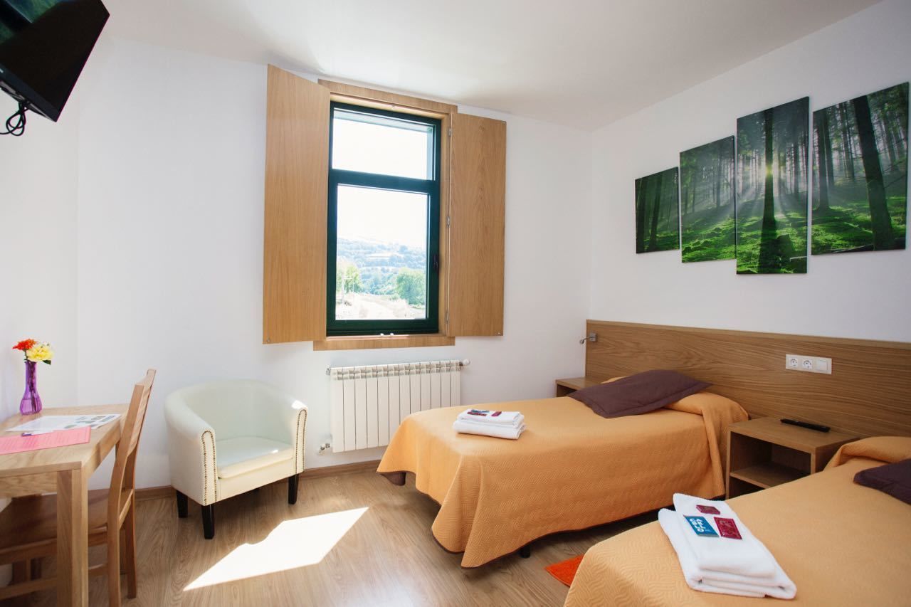 A hotel room with two beds, a wooden headboard, a window with a view, a desk with a chair, and a vase with flowers.