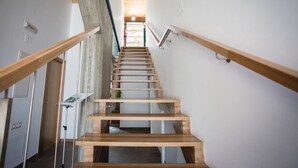 Una escalera de madera con barandas metálicas, que lleva a una habitación con un pilar de concreto y vistas al exterior.