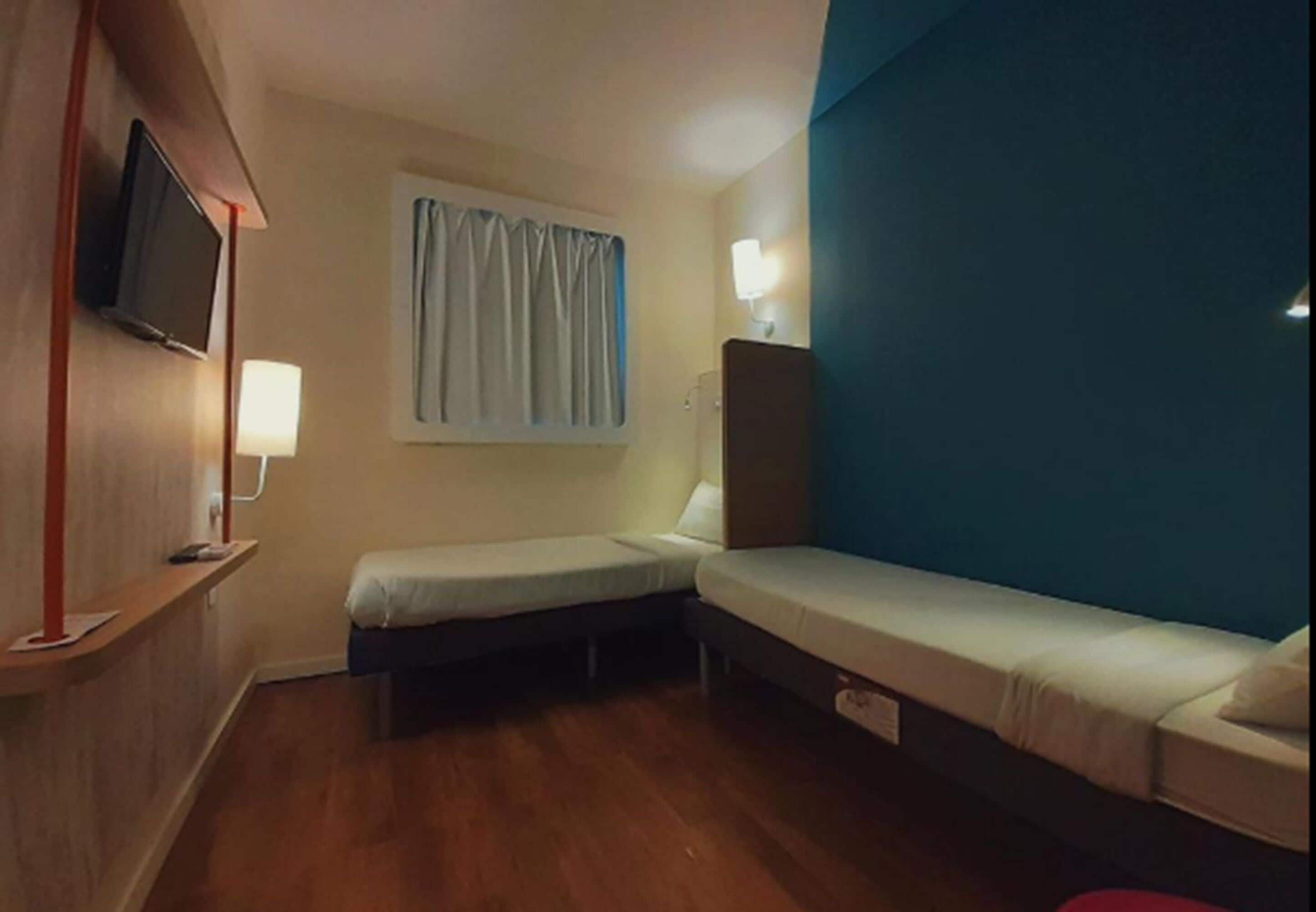 Quarto pequeno e compacto com duas camas de solteiro, televisão presa na parede, janela com cortinas e mesa de cabeceira com abajur.