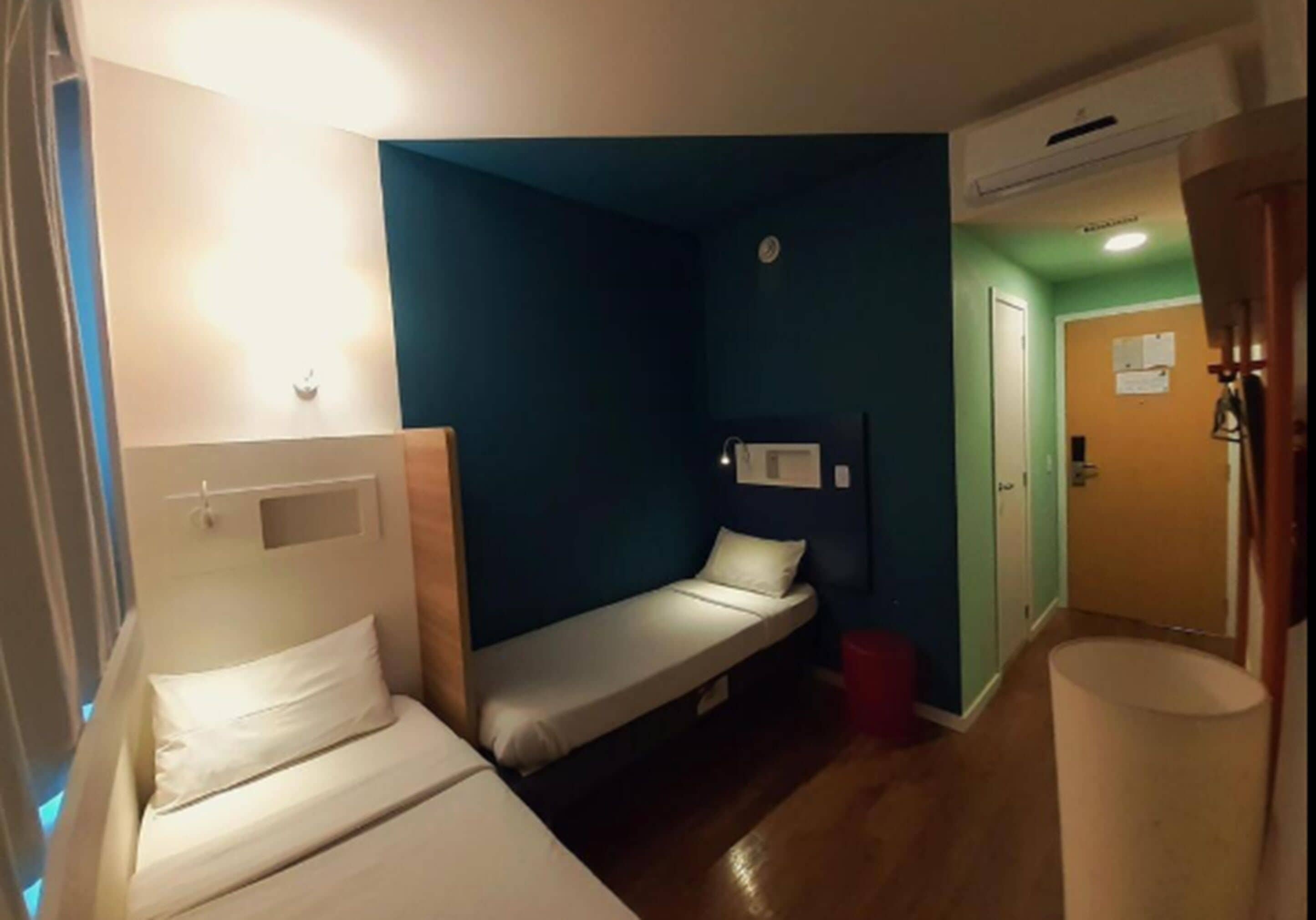 Een kleine, praktische kamer met twee éénpersoonsbedden, een houten vloer en een zichtbare deur naar een andere kamer.