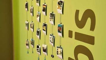 A wall with a green background and a large 'ibis' logo. There are multiple photos hanging on the wall, and a small table with a vase of flowers and a sign.