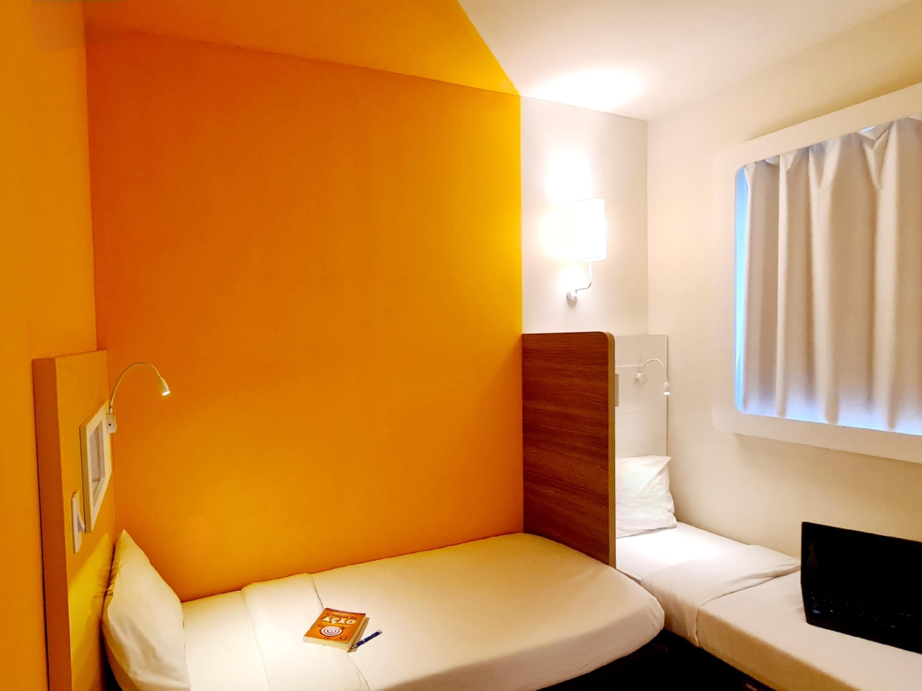 A small, single-bed room with a wooden headboard, a bedside lamp, a window with curtains, and a laptop on the bed.