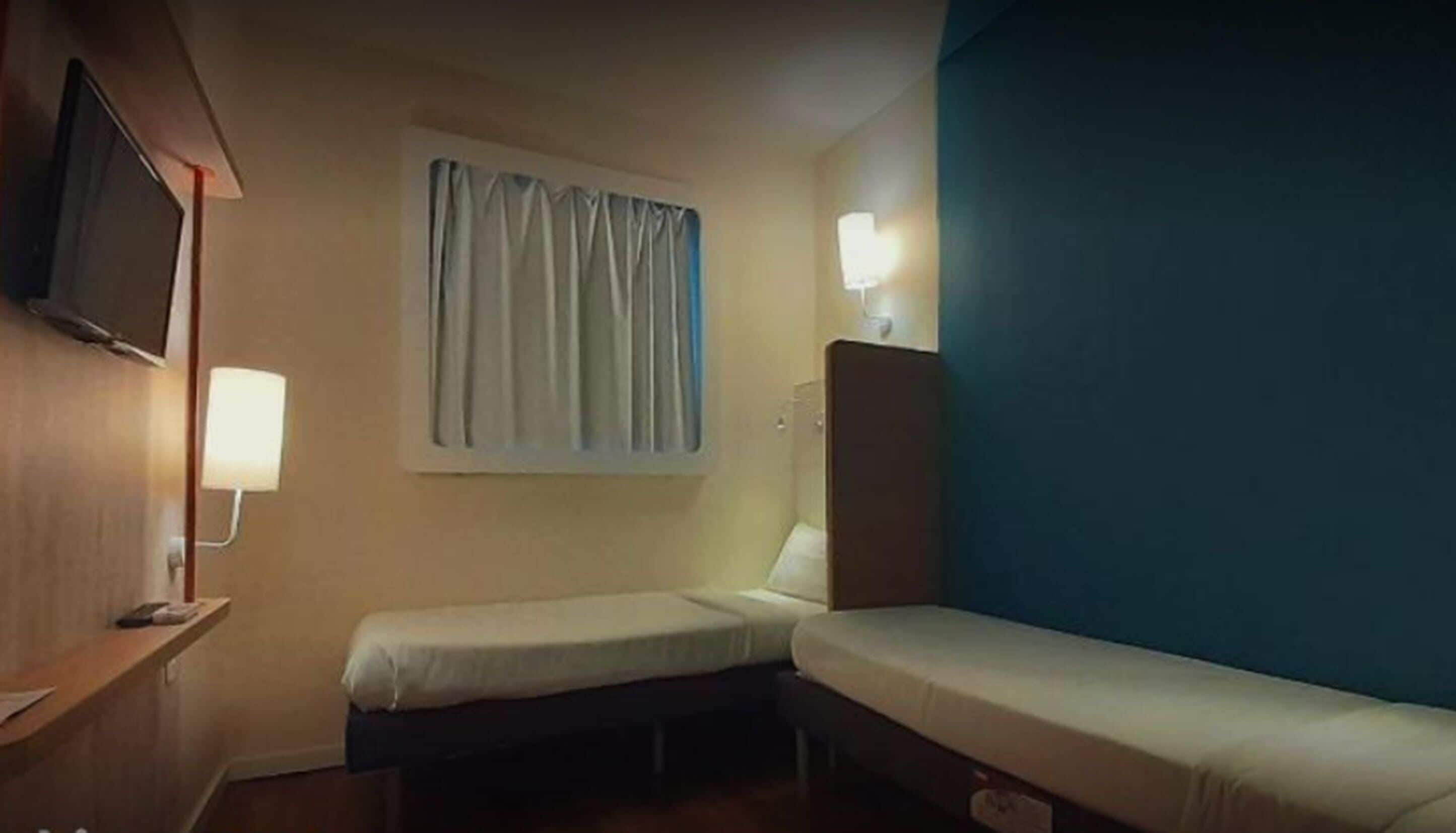 A small, single-bed room with a television on the left, a window with curtains, and a nightstand with a lamp.