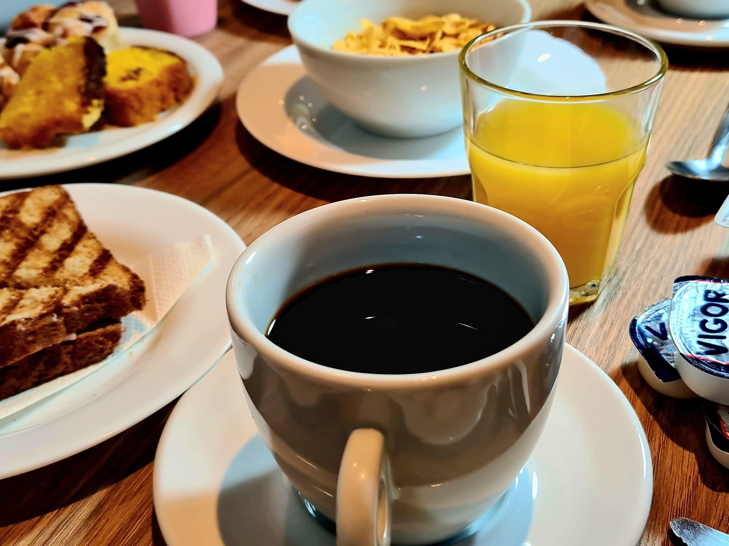 A breakfast table with a cup of black coffee, a glass of orange juice, and plates of toast and cereal.