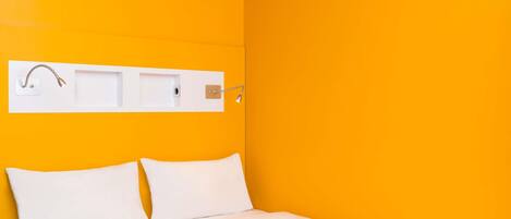 Una habitación con una cama, ropa blanca, una pared blanca y un estante blanco con un grifo y una lámpara.