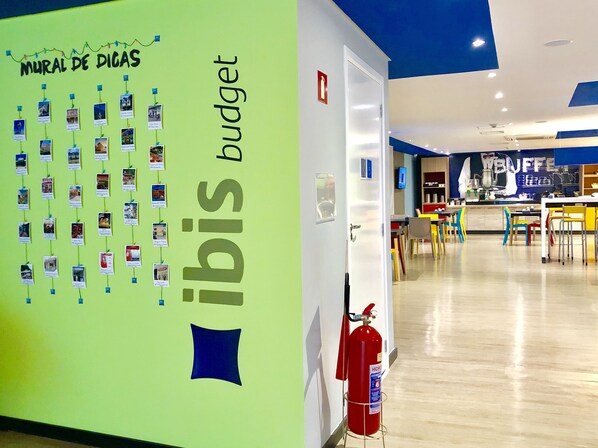 A room with a green wall featuring a 'Mural de Dicas' and 'ibis budget' signage, a fire extinguisher, a kitchen area with a 'BUFFET' sign, and a dining area with colorful chairs. - ibis budget Petropolis (Petrópolis)