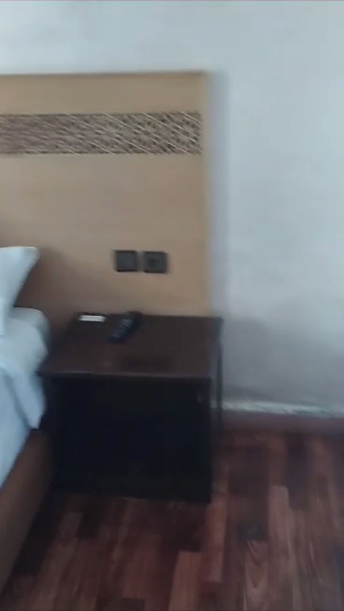 A room with a bed, a small wooden nightstand, and a wall-mounted light switch.