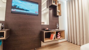 Een moderne hotelkamer met een grote televisie aan de muur, een houten console met een vaas en boeken, en een groot raam met gordijnen.