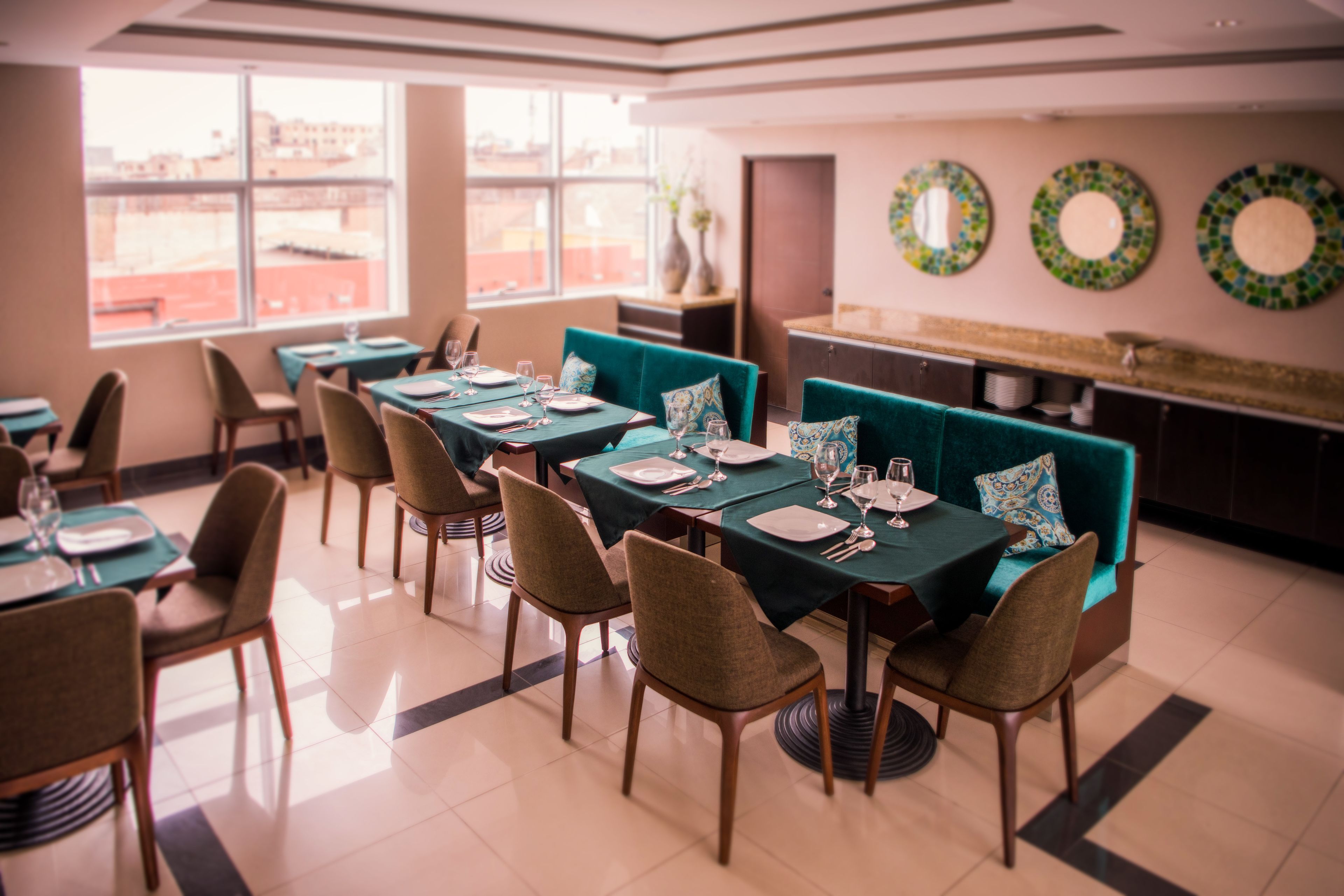 A dining area with tables set for four, teal upholstered chairs, and a view of the city through large windows.