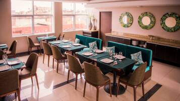 A dining area with tables set for four, teal upholstered chairs, and a view of the city through large windows.