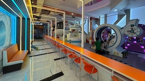 Een moderne binnenruimte met een lange oranje toonbank, meerdere oranje stoelen, en een kind dat op een futuristische attractie speelt.