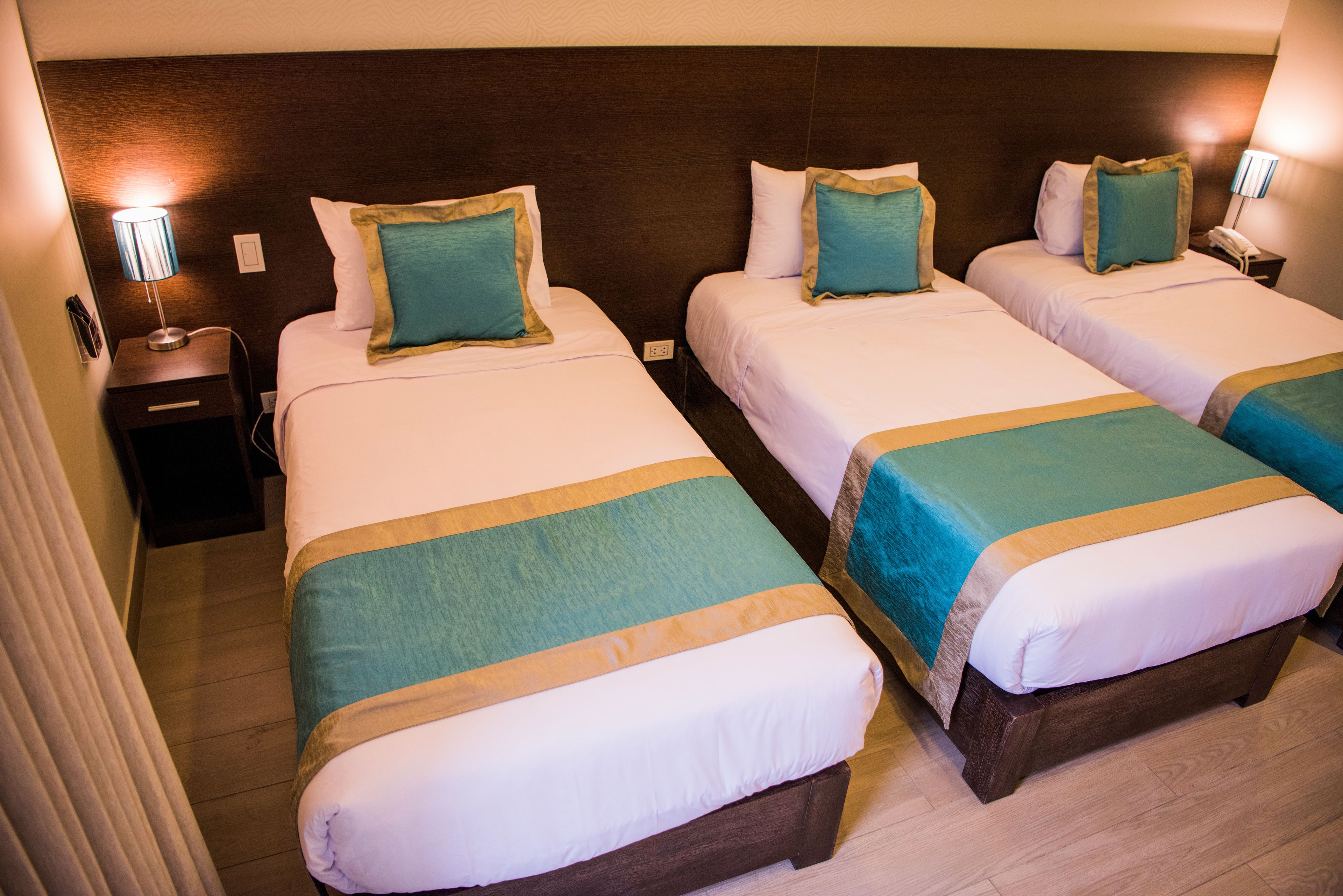 A hotel room with three single beds, each with a teal and gold bedspread, white pillows, and a wooden headboard. There is a nightstand with a lamp and a phone on each side of the beds.