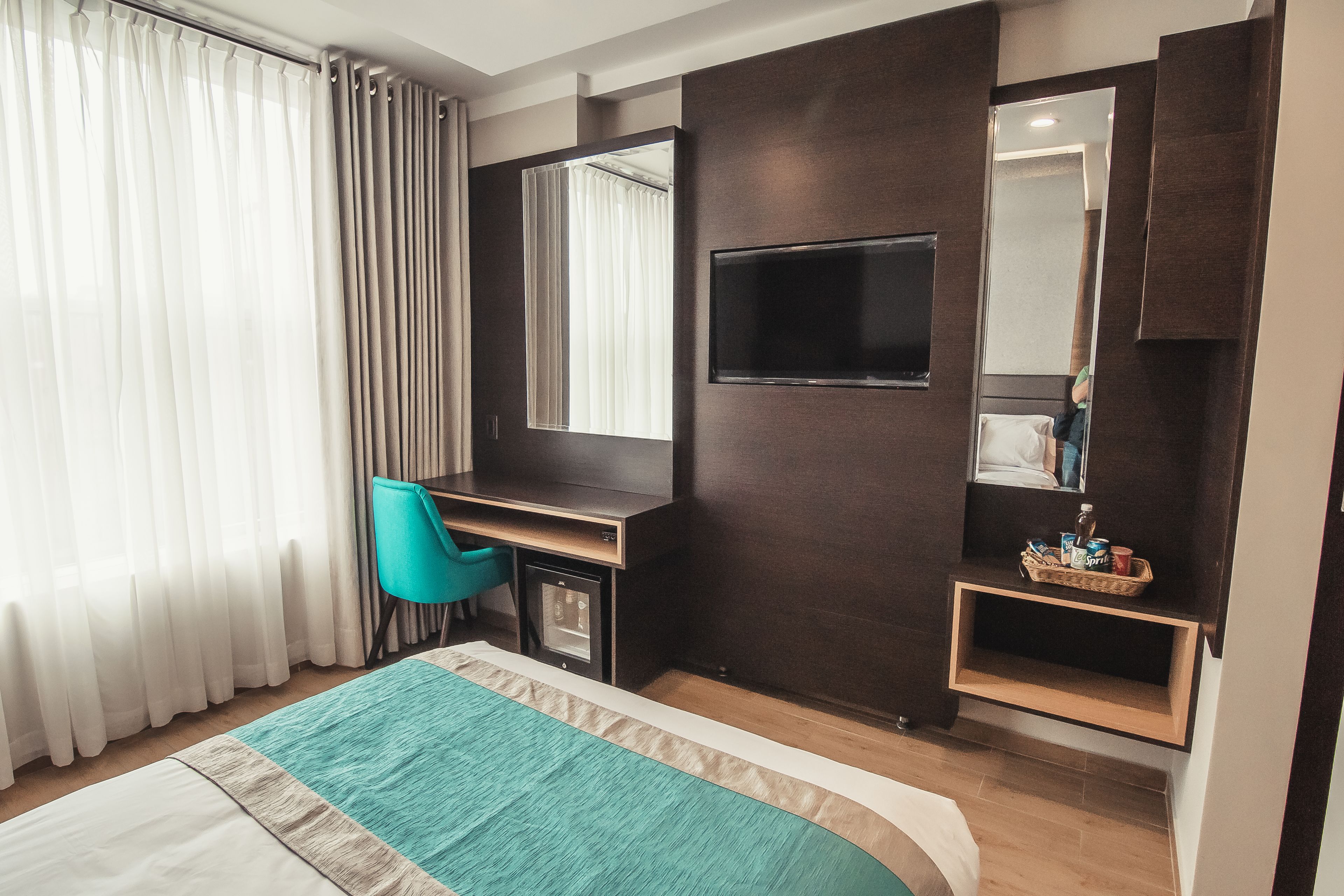 Habitación de hotel con cama, escritorio, silla, televisor, espejo y repisas con objetos.