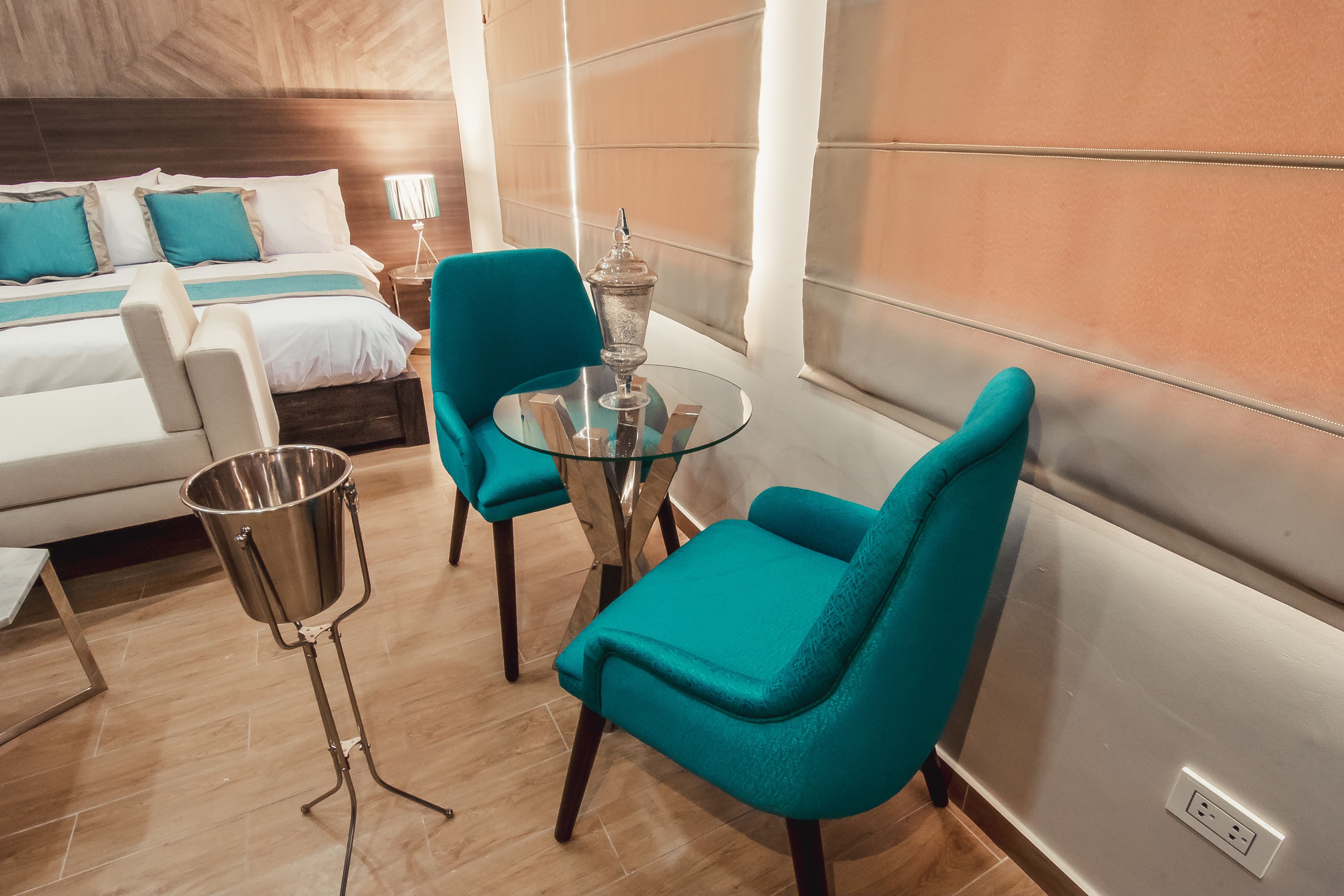 Habitación de hotel con cama, dos sillas color turquesa, una mesa de vidrio, una lámpara de metal y vistas de la ciudad.