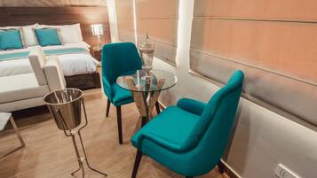 Habitación de hotel con cama, dos sillas color turquesa, una mesa de vidrio, una lámpara de metal y vistas de la ciudad.