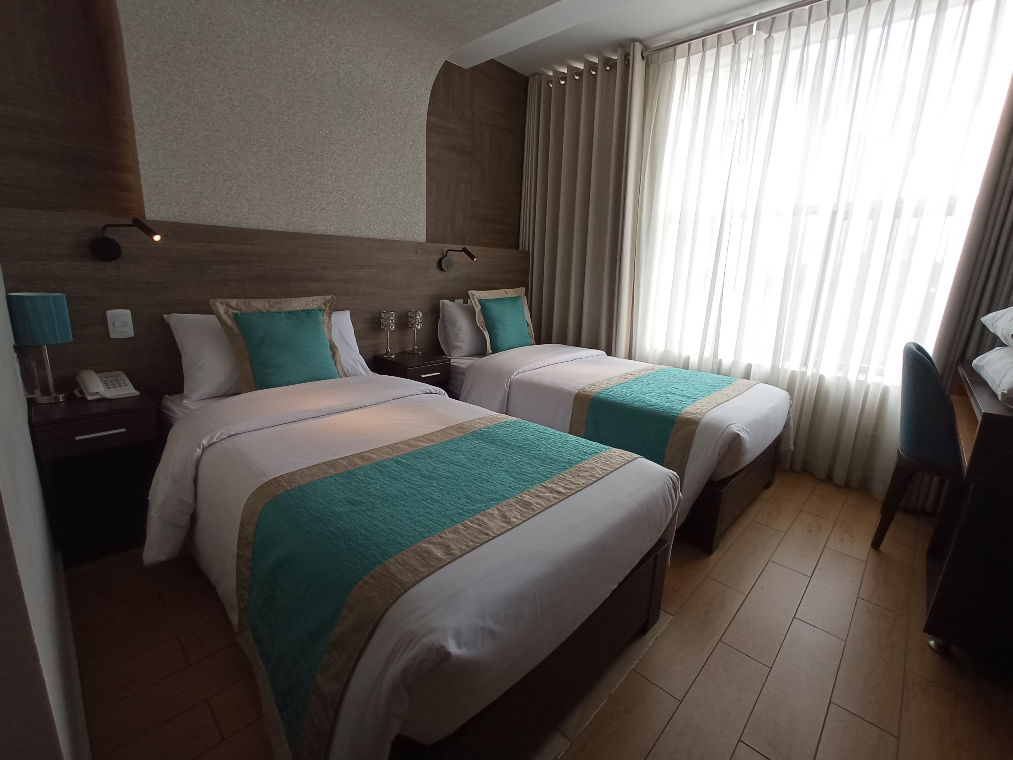 Une chambre d’hôtel avec deux lits, un bureau, une chaise, une lampe et une fenêtre avec des rideaux.