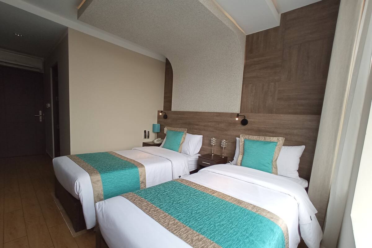 Quarto de hotel com duas camas, cabeceira de madeira, mesas de cabeceira com luminárias e uma porta à esquerda.