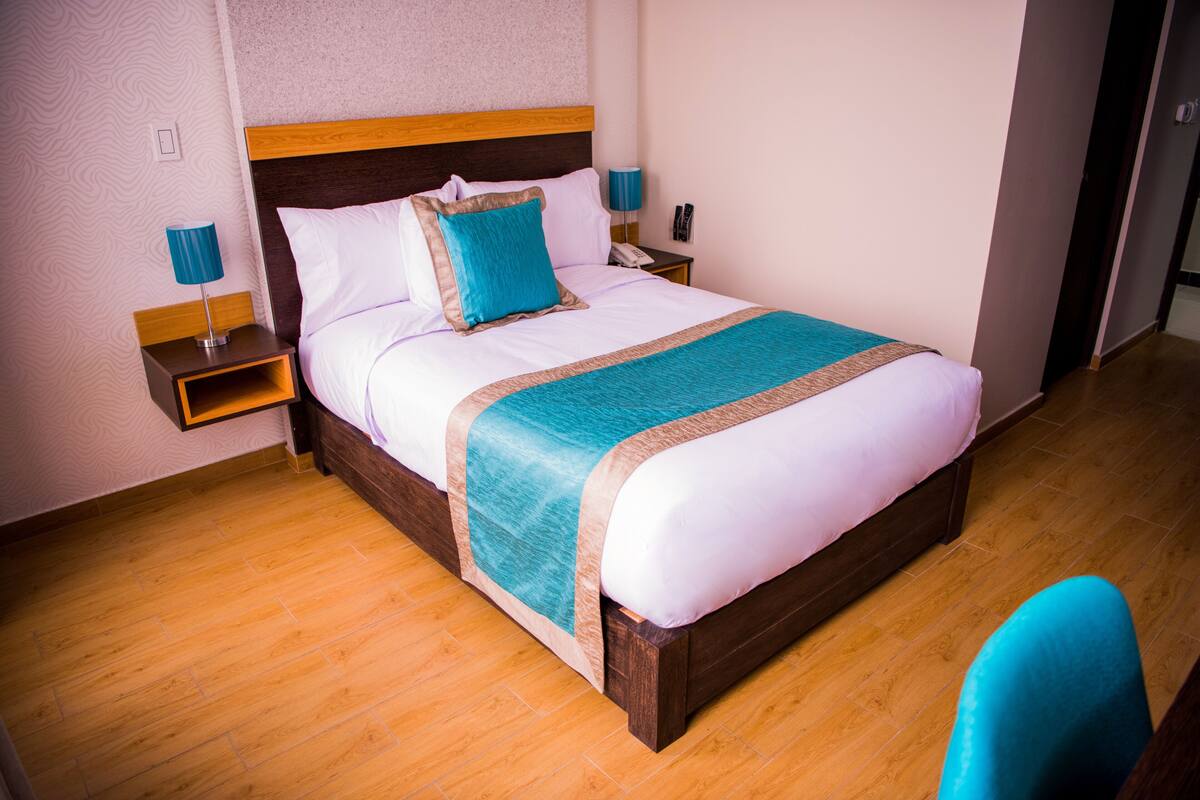 Cama bem arrumada com cabeceira de madeira, travesseiro azul e manta verde-água. Apresenta uma mesinha de cabeceira com abajur e telefone. O quarto tem piso de madeira e paredes em tons claros.