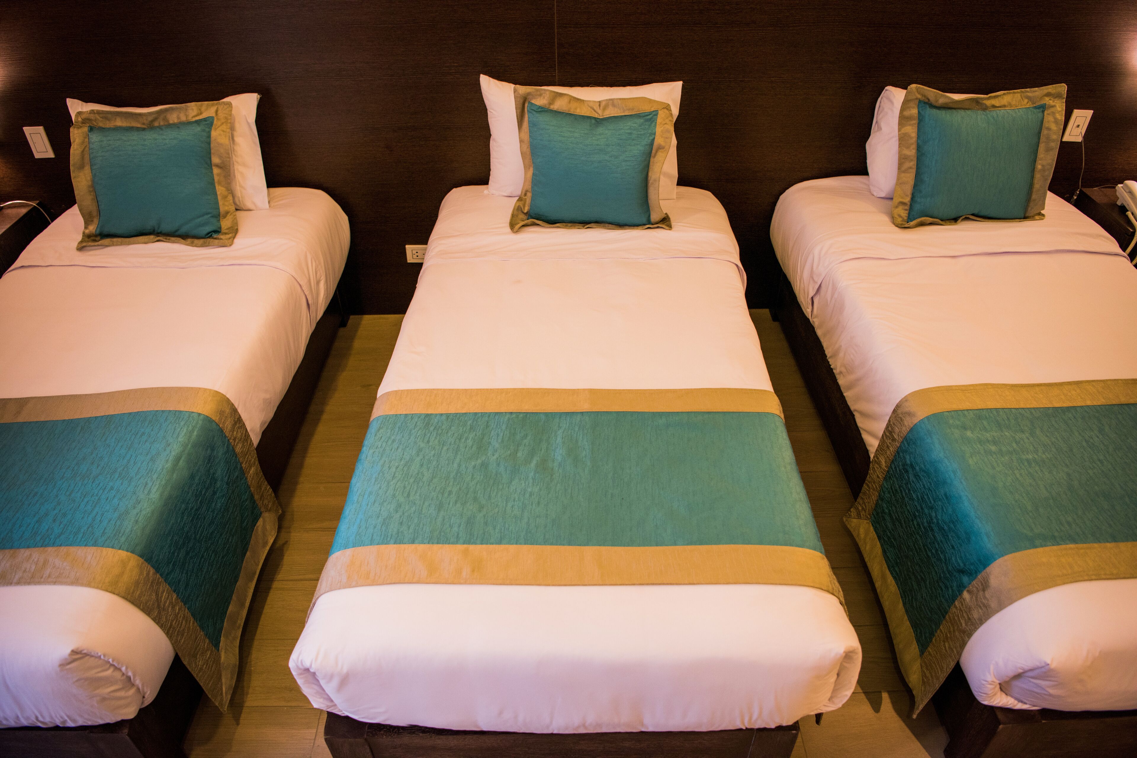 Tres camas individuales con cubrecamas verde azulado y color beige, un cabecero de madera y una mesita de noche con un teléfono.