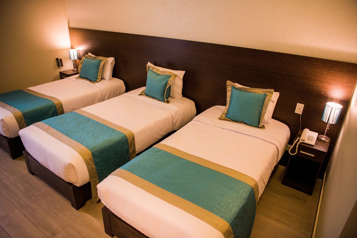 Quarto de hotel com três camas de solteiro, cada uma com colcha azul-turquesa e bege, e cabeceira de madeira. Há uma mesa de cabeceira com abajur e telefone ao lado direito.
