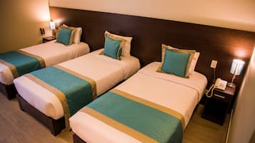 Habitación de hotel con tres camas individuales, cada una con colcha verde aguamarina y color crema, y cabecero de madera. Se observa una mesita de noche con lámpara y teléfono a la derecha.