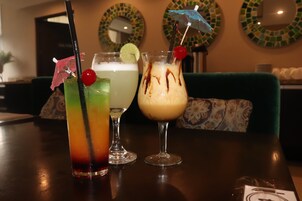 Trois cocktails sont posés sur une table, avec un canapé vert et des miroirs décoratifs en arrière-plan.
