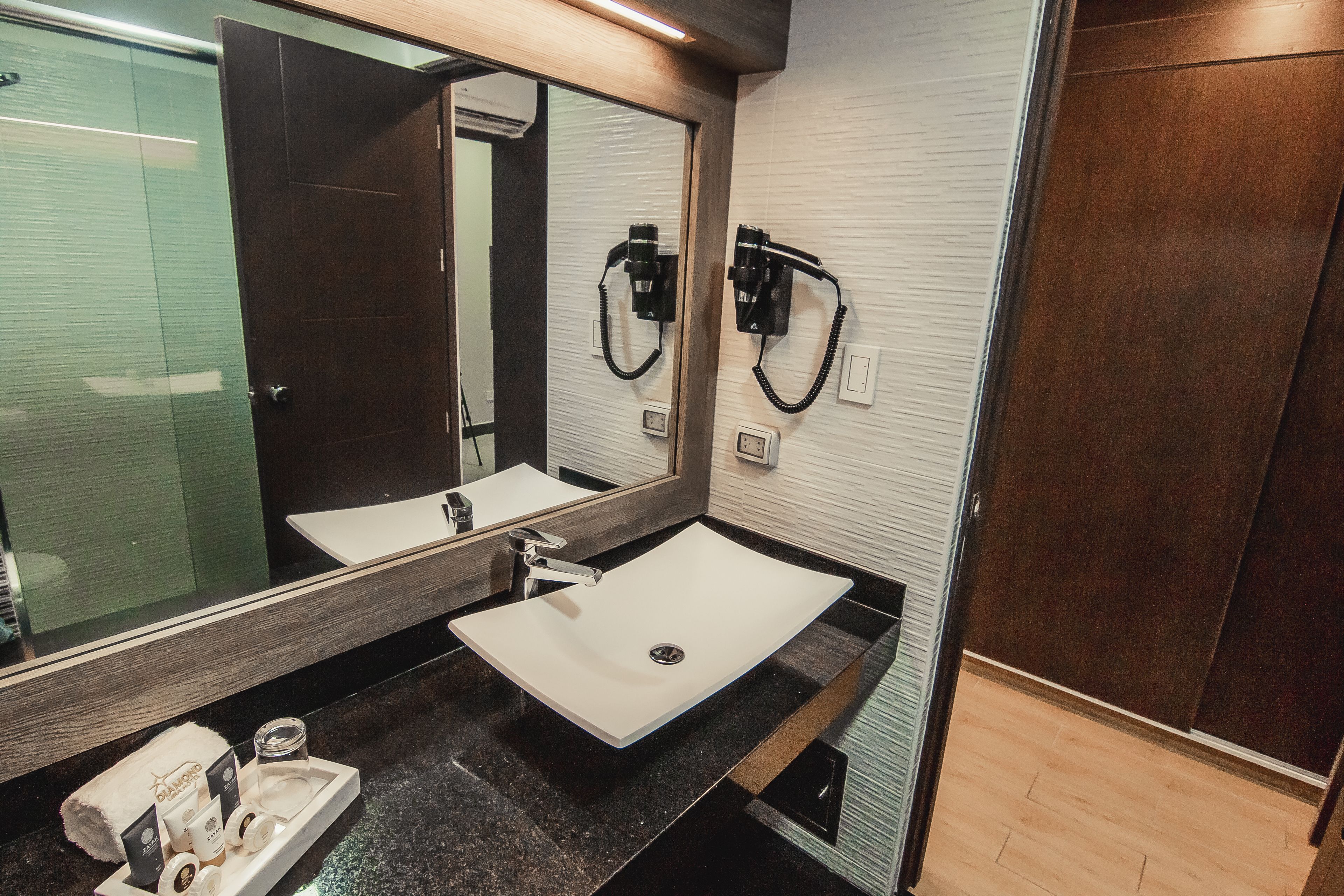 A modern bathroom with a large mirror, a white sink, and a black countertop. There are two hair dryers mounted on the wall, and a towel rack with a towel is visible.