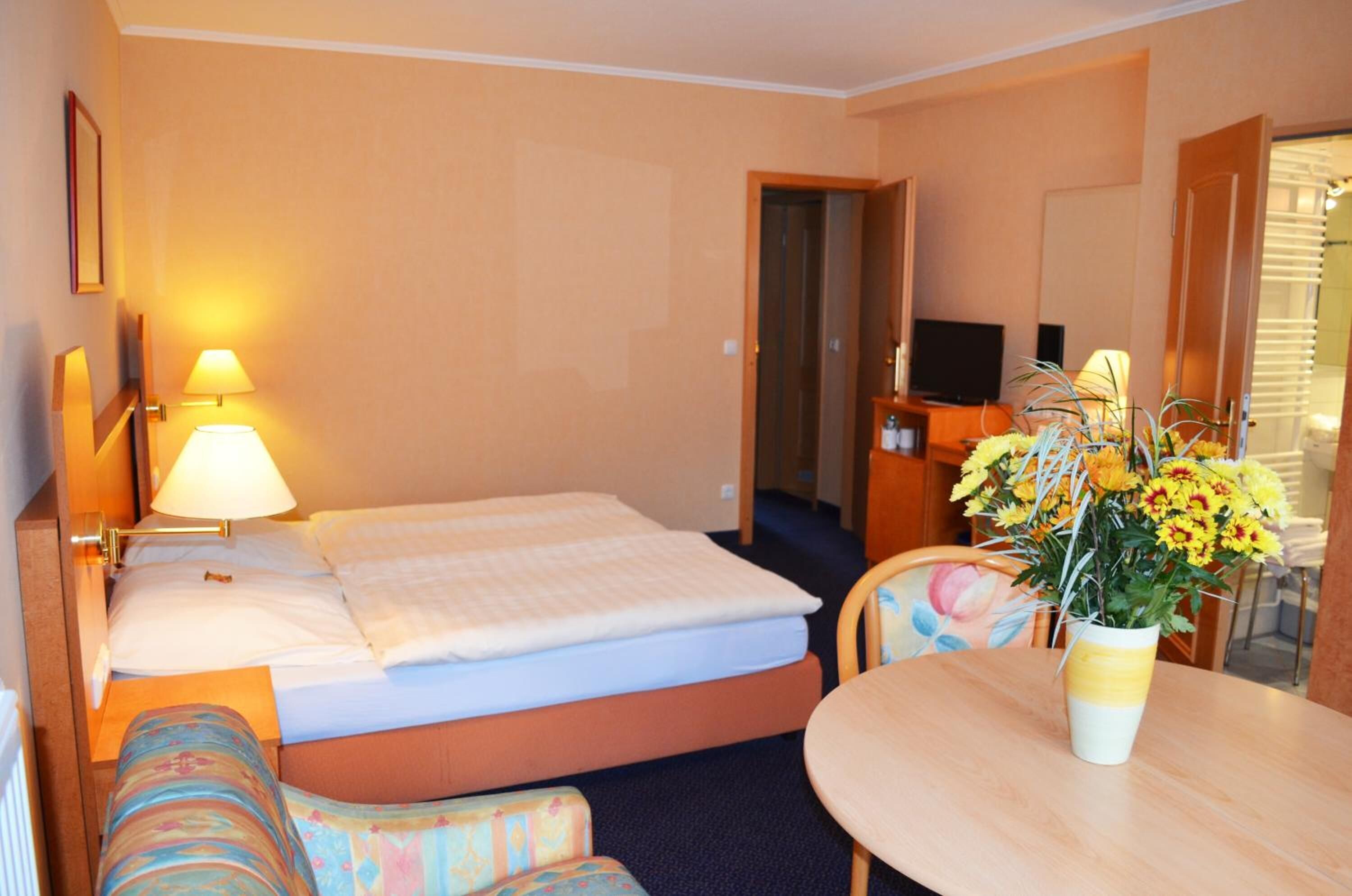 A hotel room with a bed, a bedside lamp, a small table with a vase of flowers, and a television in the corner.