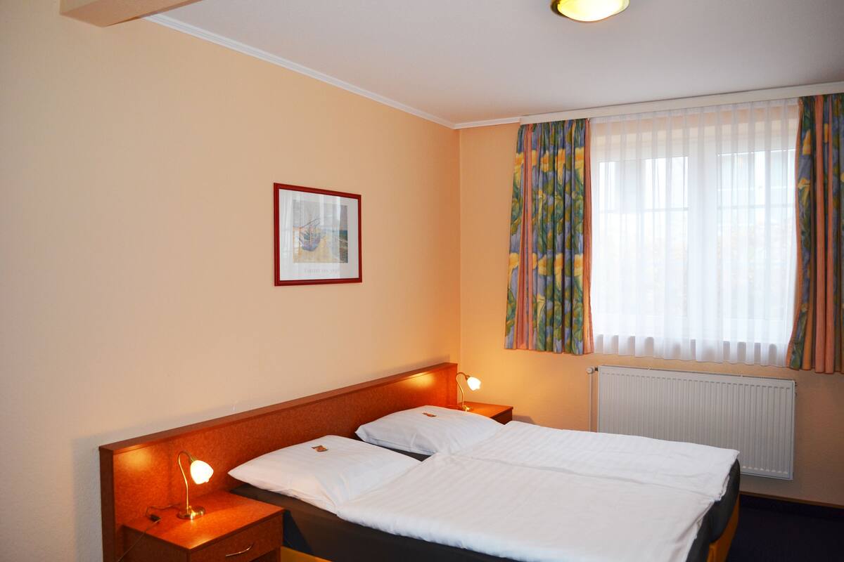 Une chambre d’hôtel avec un lit double, deux lampes de chevet, un tableau encadré au mur et une fenêtre avec des rideaux.