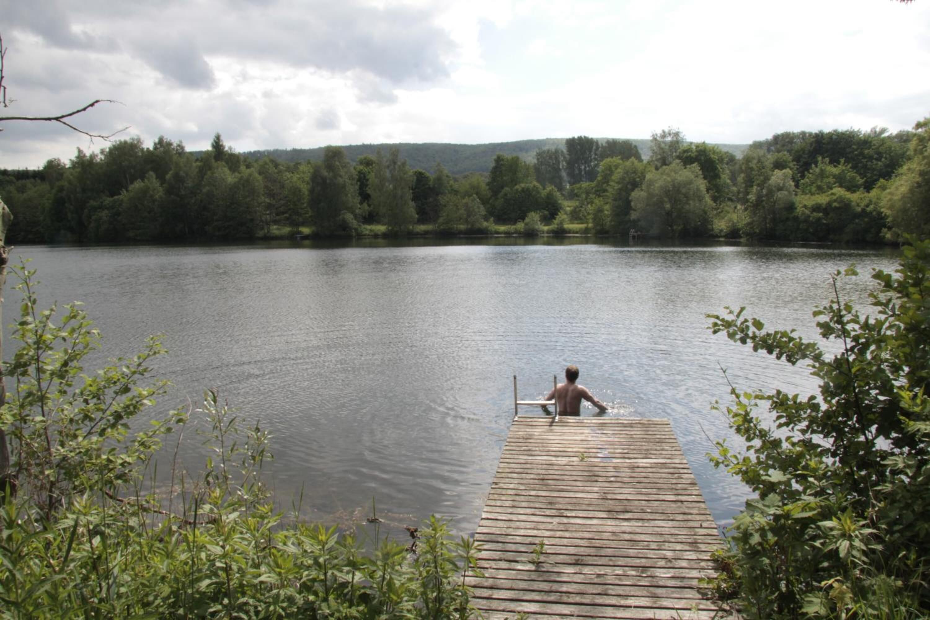 Une jetée en bois s’étendant sur un lac paisible, entouré d’une végétation luxuriante et avec des collines en arrière-plan.