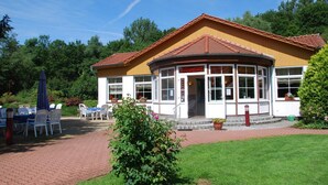 A building with a curved roof and large windows, surrounded by a garden with tables and chairs.