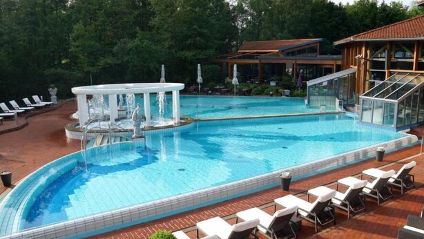 A swimming pool with a fountain, surrounded by lounge chairs and a covered area with a view of trees.