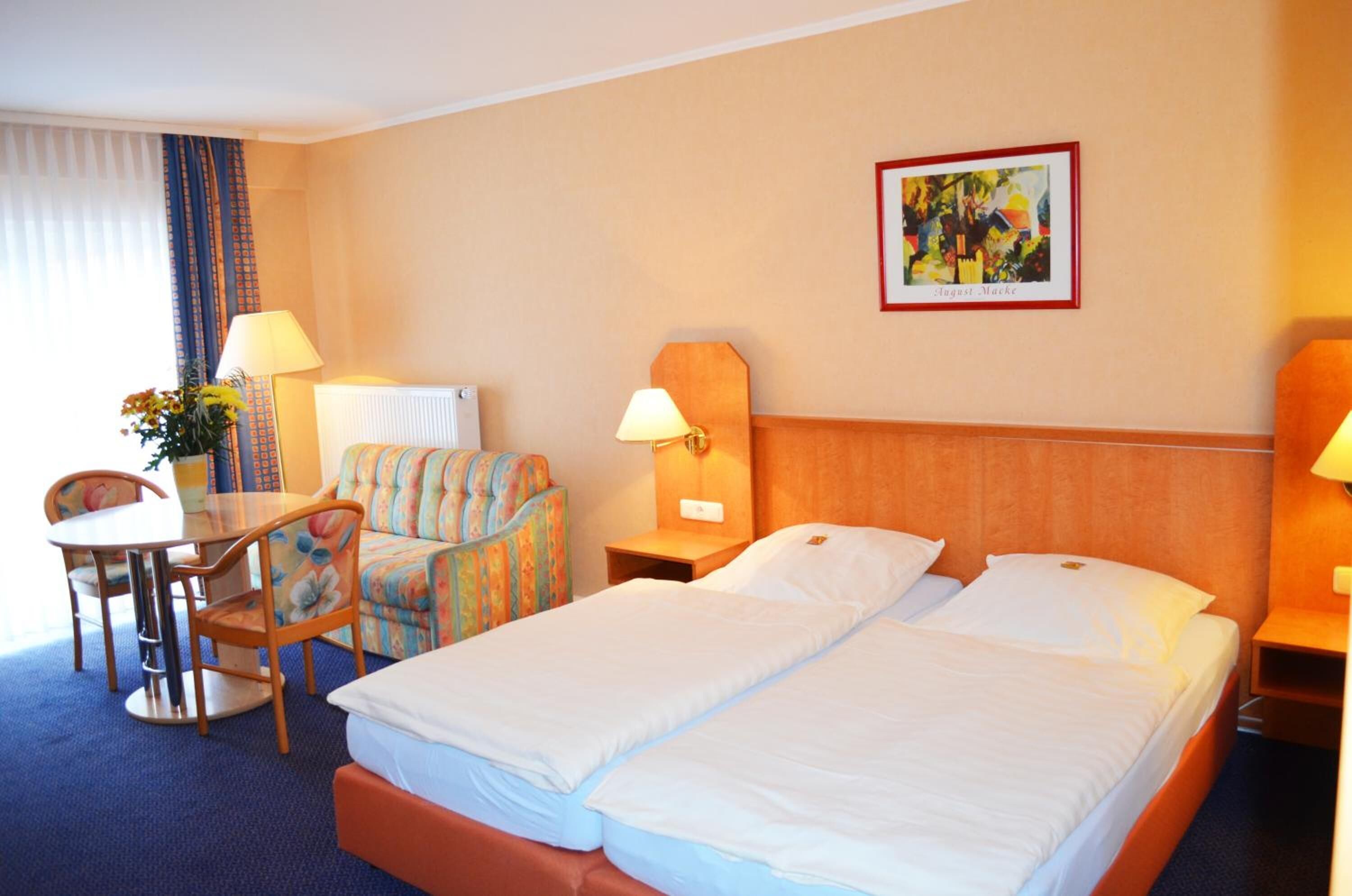 Ein Hotelzimmer mit zwei Betten, einem Schreibtisch, einem Stuhl, einer Lampe und einem gerahmten Bild an der Wand.
