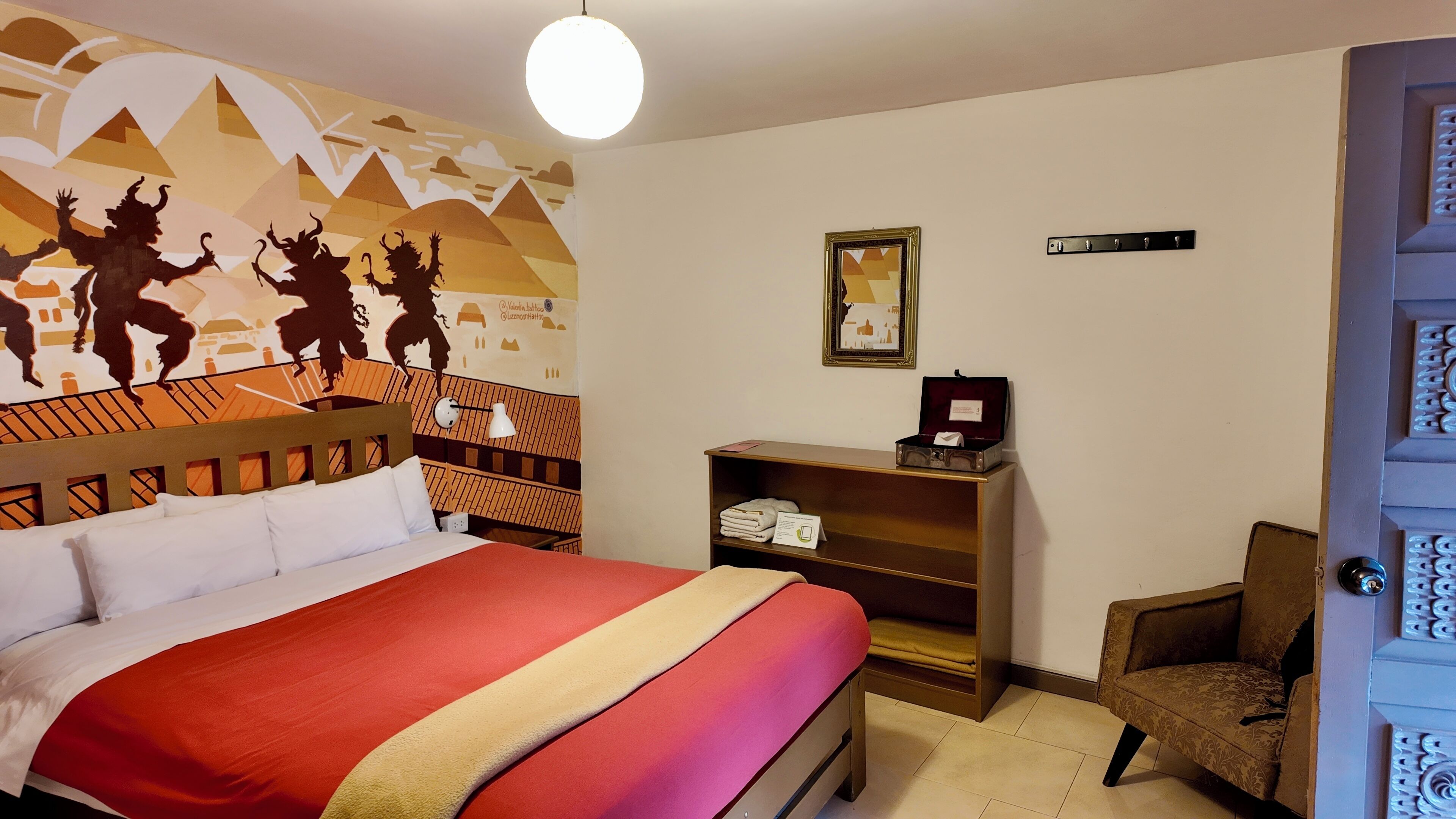 Quarto de hotel com uma cama, mesa de cabeceira, cadeira, parede com mural, prateleira com objetos e porta com painel decorativo.