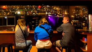 Three people seated at a bar with a city view through a window. - Pariwana Hostel Cusco - Adults only (Cusco)