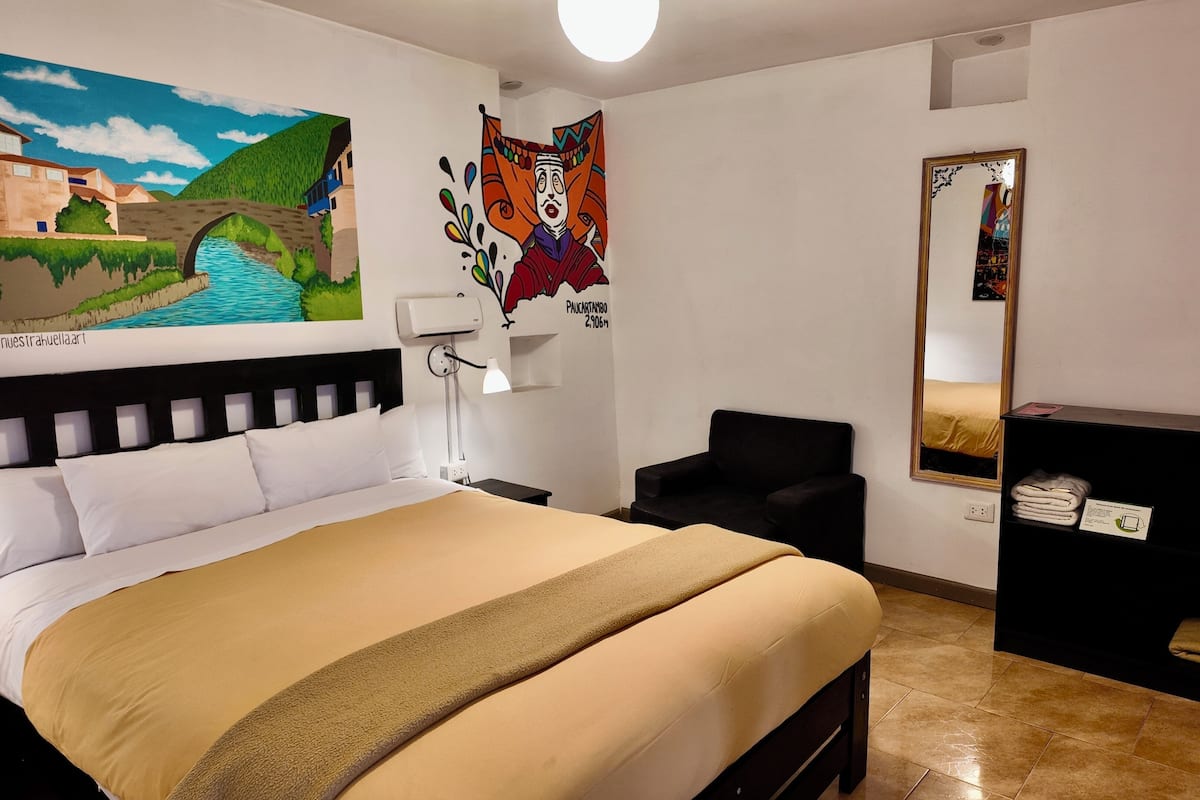 Habitación de hotel con una cama, una silla, un escritorio, un espejo y dos cuadros en la pared.