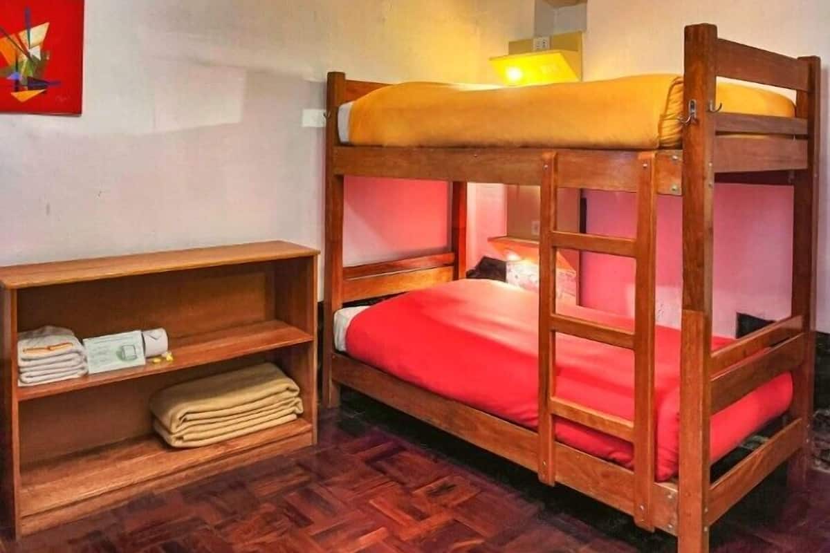 Una cama litera con colchón rojo, un estante de madera con toallas dobladas y una lámpara en la cama superior.