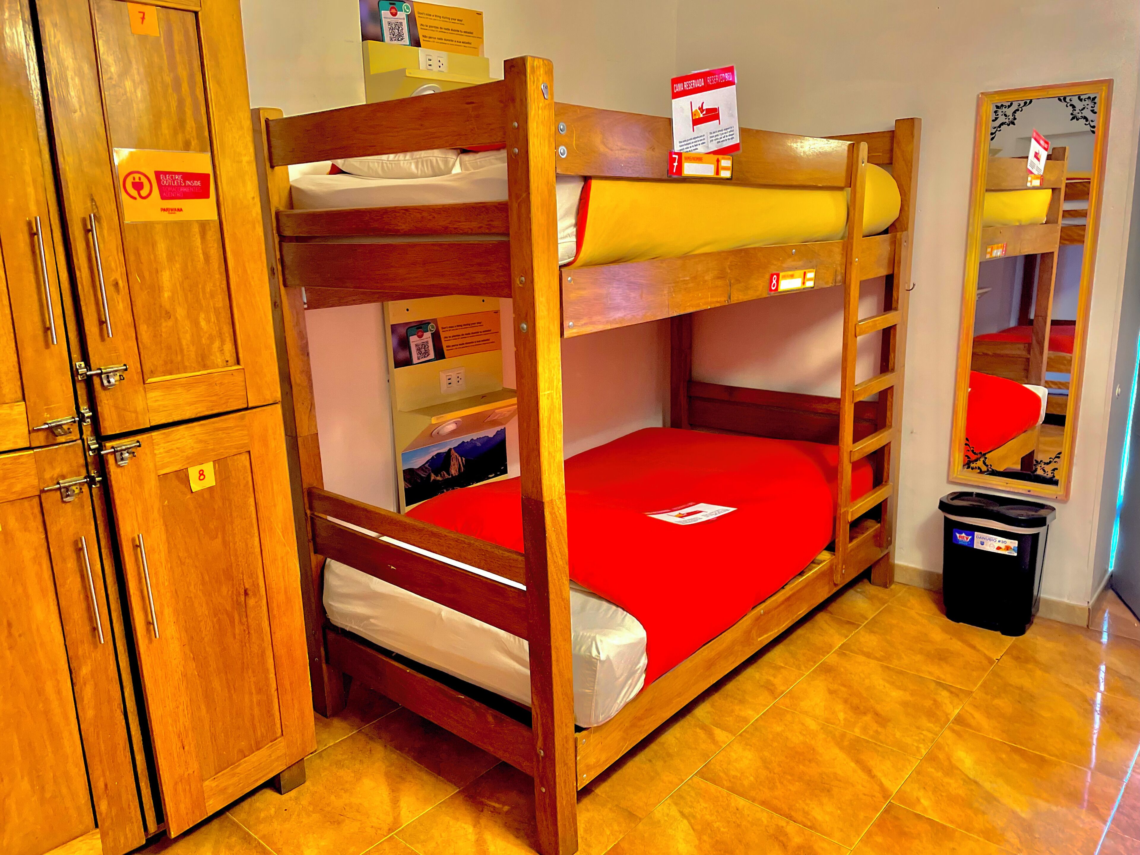 Una cama litera con colchón rojo, literas de madera, un armario con un código QR y un cubo de basura.