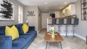A modern living room with a blue sofa, a wooden coffee table, and a kitchen area with a bar stool set. - Elliot Oliver - 2 Bedroom Garden Apartment With Parking (Cheltenham)