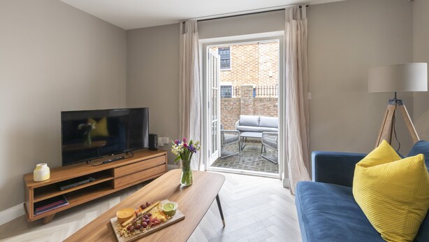 A modern living room with a flat-screen TV, wooden entertainment unit, a coffee table with snacks, a sofa with a yellow pillow, and a balcony with outdoor seating. - Elliot Oliver - 2 Bedroom Garden Apartment With Parking (Cheltenham)