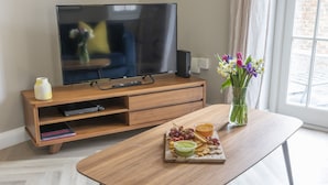 A modern living room with a wooden entertainment unit, a flat-screen TV, a glass vase with flowers, and a wooden coffee table with a board of snacks. - Elliot Oliver - 2 Bedroom Garden Apartment With Parking (Cheltenham)