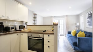 A modern kitchen with white cabinets, a microwave, and a toaster. There is a blue sofa with yellow cushions in the living area. - Elliot Oliver - 2 Bedroom Garden Apartment With Parking (Cheltenham)