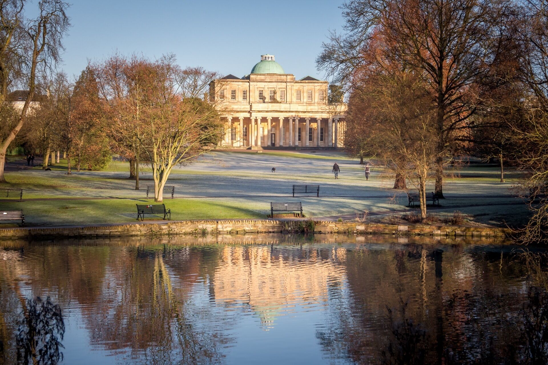 A large, classical building with a dome, surrounded by a park with benches and a pond reflecting the structure.