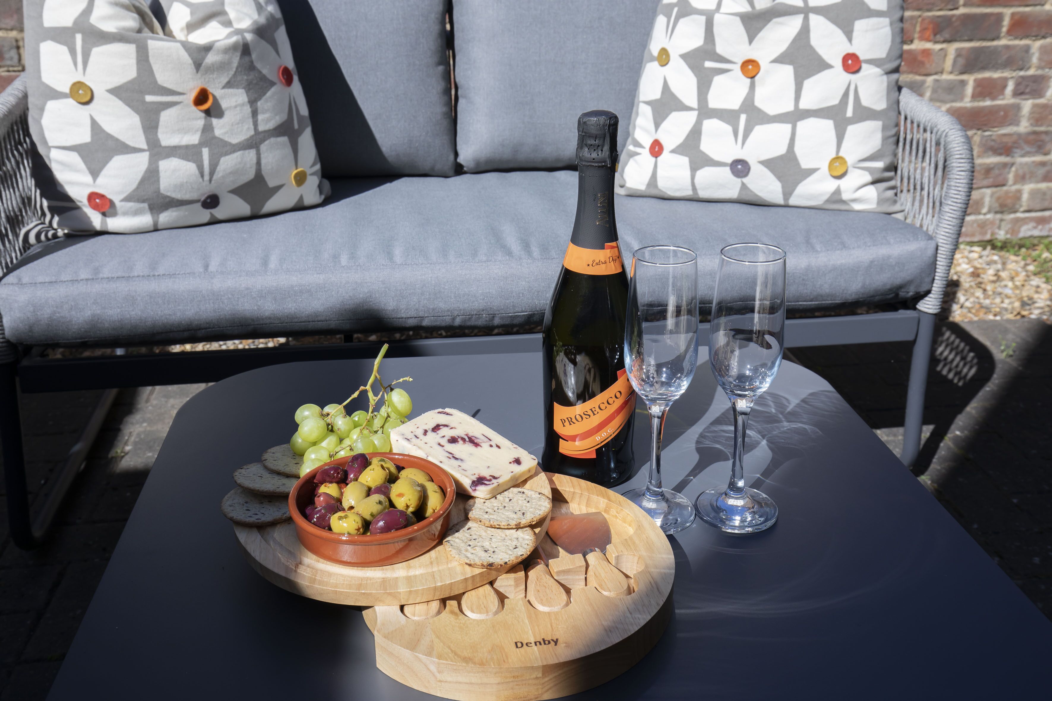 Une table avec une bouteille de prosecco, deux verres, un bol d’olives, des raisins et du fromage, le tout disposé sur une planche en bois.